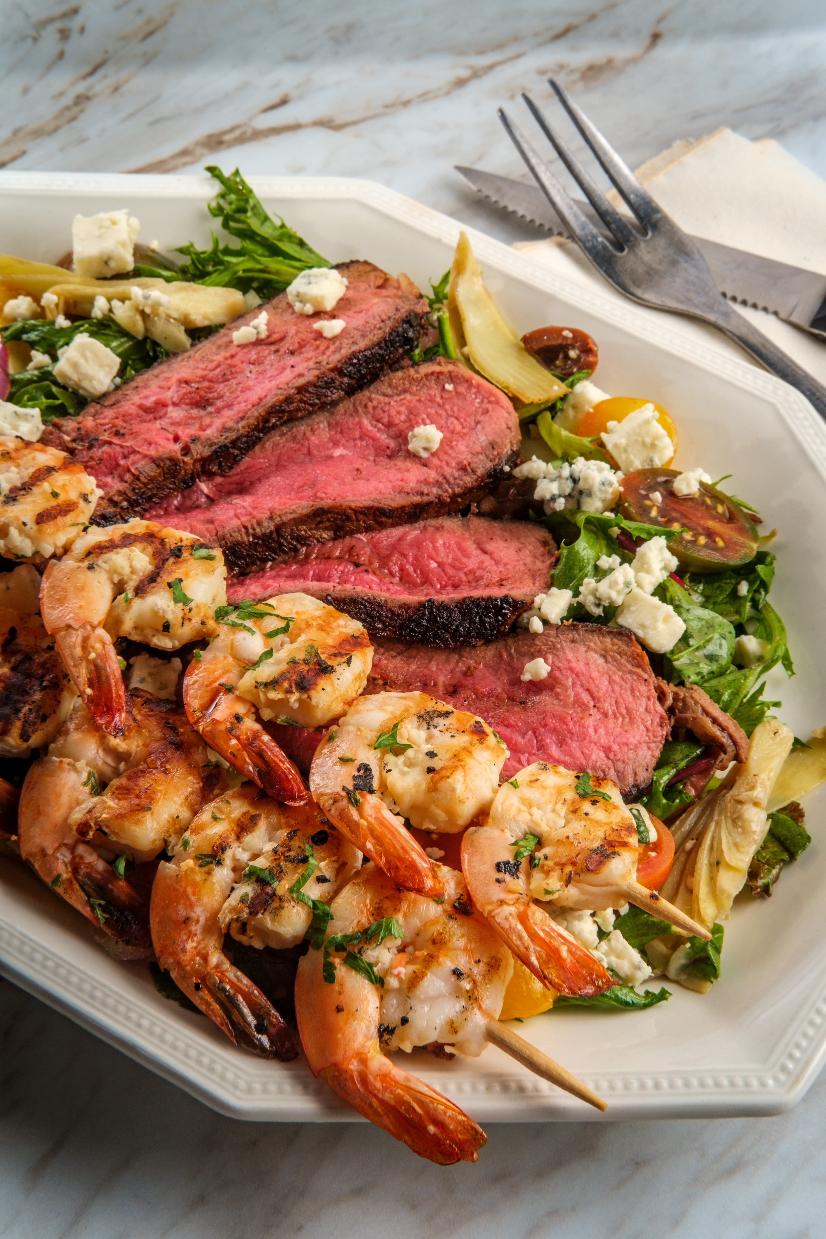 Sliced medium-rare steak and grilled prawn skewers on a platter with salad leaves
